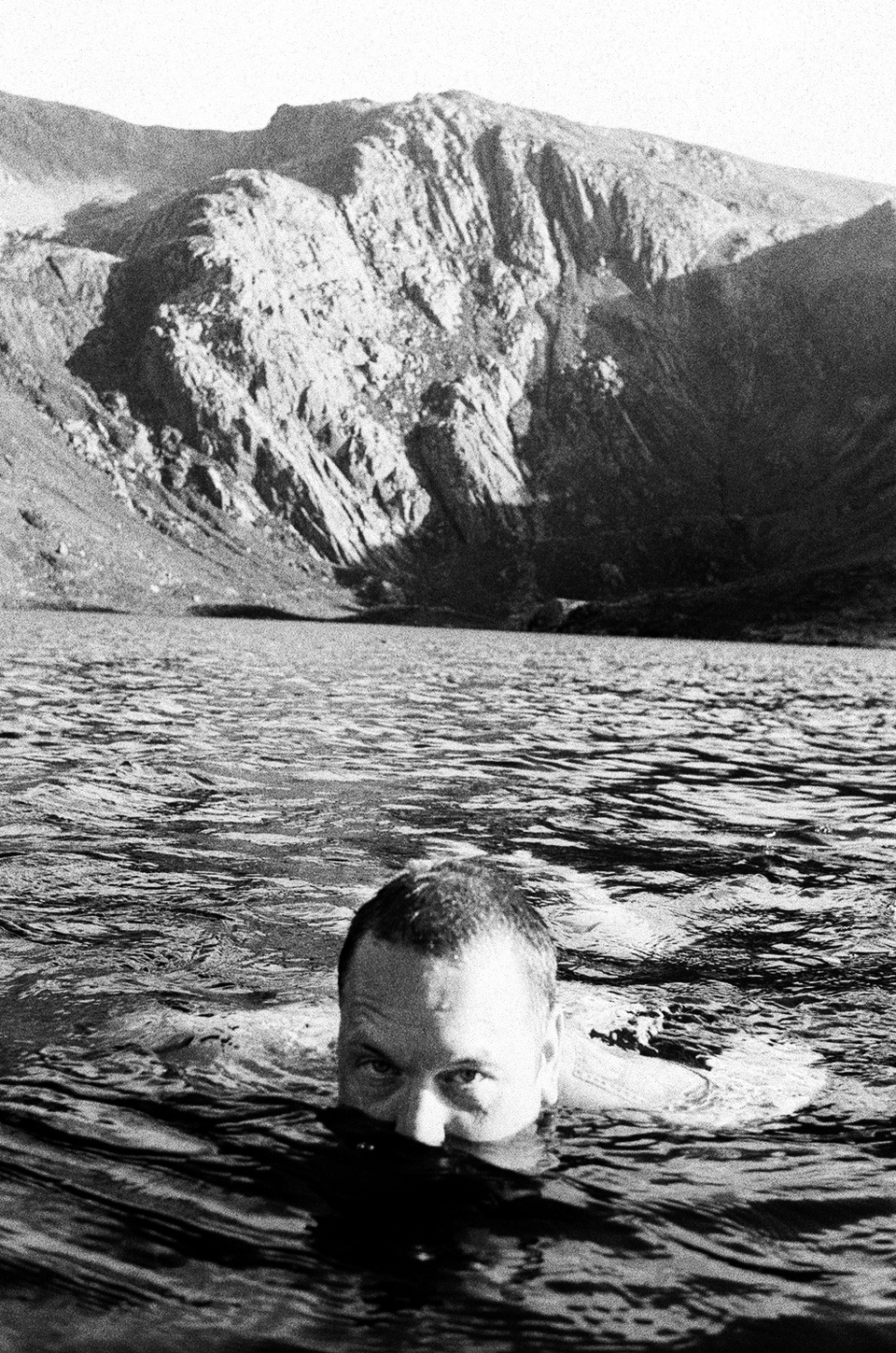 Black and white photo of a person partially submerged in a lake, with only their eyes and forehead visible above the water. Rocky, mountainous terrain rises in the background under bright sunlight.